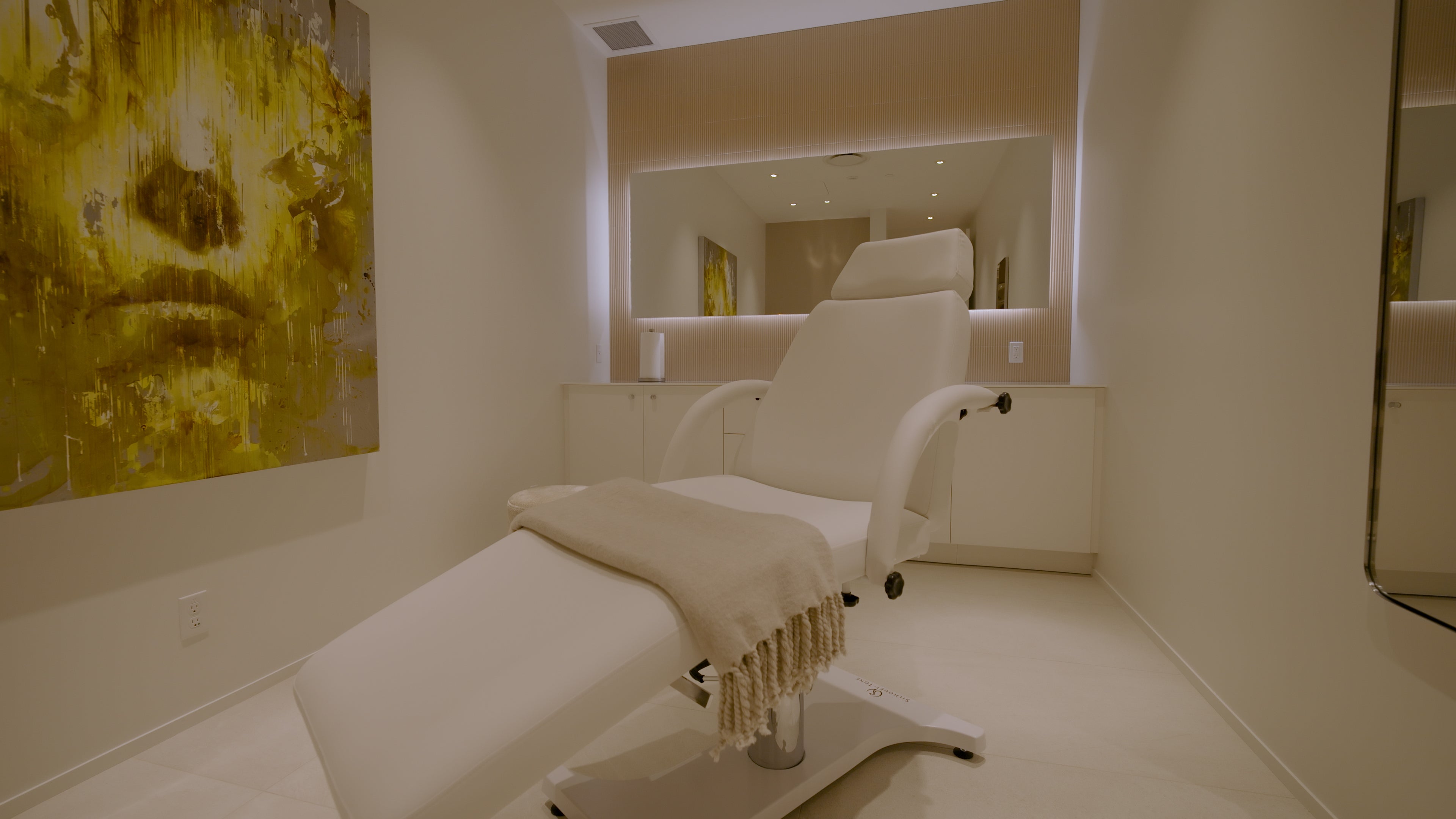 Beige massage chair in a spa-like room with a large mirror and abstract art on the wall.